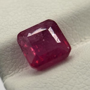 May include: A vibrant, square-cut ruby gemstone with a deep red hue. The gemstone is set against a white surface, showcasing its facets and brilliance. The ruby's color is rich and saturated, reflecting light.
