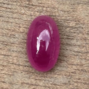 Op de afbeelding: Een gepolijste, ovaalvormige robijn edelsteen. De edelsteen heeft een diepe, verzadigde roze-rode kleur met een glad, reflecterend oppervlak. De edelsteen ligt op een houten achtergrond.