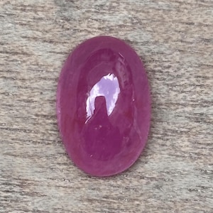 Pode incluir: Uma pedra preciosa de rubi polida, em forma oval. A gema é de uma cor vermelho-rosada profunda e saturada, com uma superfície brilhante. A pedra está sobre um fundo de madeira.