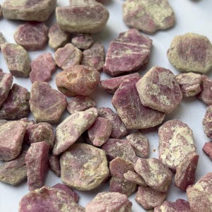 May include: A collection of raw, unpolished ruby gemstones in various shapes and sizes. The stones feature a mix of deep magenta, pink, and muted off-white or beige mineral inclusions, displaying natural, jagged edges and uneven, textured surfaces.