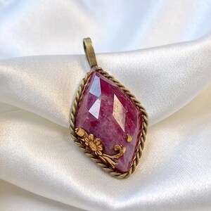 May include: A diamond-shaped pendant featuring a faceted deep pink gemstone set in a twisted antique brass frame. The setting includes a small decorative brass floral accent on the lower left side and a simple bail at the top for hanging.