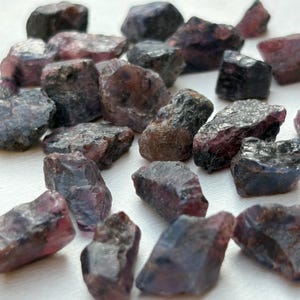 May include: A collection of rough-cut gemstones in shades of deep purple, burgundy, and black. The stones have a natural, unpolished appearance, with visible textures and variations in color. The arrangement is scattered, showcasing the individual shapes and hues of each stone.