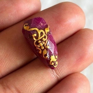 May include: A deep red gemstone with gold filigree design. The gemstone is held in a hand, showcasing its polished surface and intricate golden patterns. The gold design appears to be hand-painted, adding to the unique character of the stone.