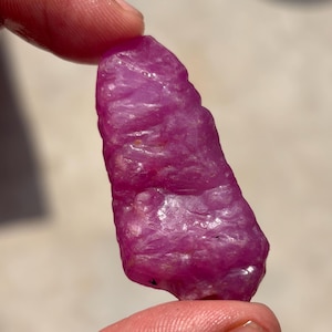Puede incluir: Una piedra preciosa en bruto y sin tallar con un tono magenta rosado vibrante. La piedra presenta una forma irregular y alargada con una superficie cristalina texturizada que capta la luz, mostrando facetas naturales y áreas translúcidas en toda su estructura.