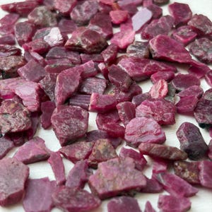 May include: A collection of raw, unpolished ruby gemstones in various shades of deep pink and magenta. The stones are irregular in shape, featuring rough, textured surfaces and opaque to translucent qualities, scattered across the frame.