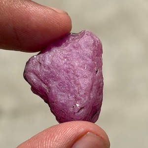 Puede incluir: Una piedra preciosa en bruto y sin pulir con un tono magenta y rosa intenso. La piedra presenta una superficie naturalmente texturizada e irregular con facetas cristalinas visibles y pequeños puntos reflectantes.