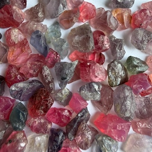 Puede incluir: Una colección de piedras preciosas en bruto y sin pulir en varios tonos de rosa, rojo, morado y gris. Estos pequeños fragmentos minerales irregulares presentan texturas translúcidas y opacas con facetas naturales y bordes dentados dispersos.