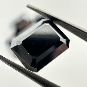 Peut inclure: Une pierre précieuse noire taille émeraude avec une surface hautement polie et réfléchissante. La pierre présente des bords biseautés nets et une forme rectangulaire, maintenue par une pince métallique sur un fond clair et uni.