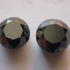 Puede incluir: Dos gemas negras redondas y facetadas. Las gemas tienen un acabado brillante y reflejan la luz, mostrando sus múltiples facetas. Las piedras están sobre un fondo liso y claro, resaltando su color oscuro y forma.