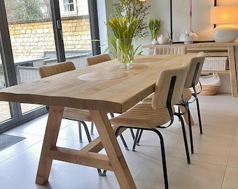May include: A rectangular light wood dining table with a trestle base and four upholstered chairs featuring black metal frames. The table is set with circular placemats and a glass vase containing yellow tulips. The word WAX is in the corner.