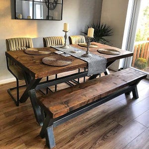 May include: A rectangular dining table with a thick dark wood top and black metal X-frame legs, accompanied by a matching wooden bench and three upholstered chairs. A grey runner, woven placemats, and two tall candle holders sit on the table.