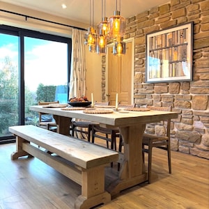 May include: A light-coloured wooden dining table with a matching bench and chairs, featuring woven placemats, two taper candles, and a bowl of fruit. Above the table hangs a cluster of five glass pendant lights with warm glowing bulbs.
