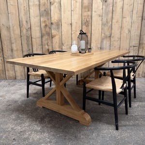 May include: A rectangular light-toned wooden dining table with a sturdy X-shaped trestle base, accompanied by four black wooden chairs featuring curved backrests and woven seats. A white pineapple-shaped object and a black lantern sit on top.