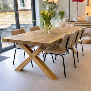 May include: A rectangular wooden dining table with a thick top and a cross-legged base, accompanied by six chairs featuring beige upholstered seats and black metal frames. A clear glass vase with yellow flowers sits in the centre of the table.