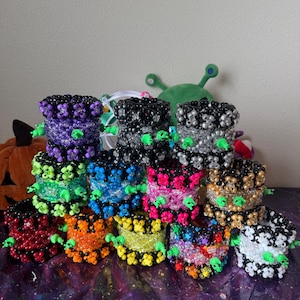 May include: A collection of twelve cylindrical beaded cuffs arranged in a group. Each cuff is constructed from small, colorful plastic beads and features small, bright green plastic alien-shaped charms attached to the exterior surface.