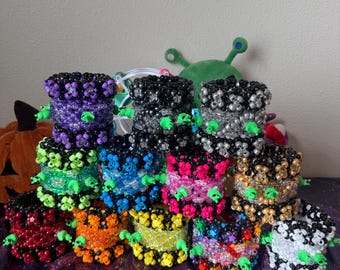 May include: A collection of twelve cylindrical beaded cuffs arranged in a group. Each cuff is constructed from small, colorful plastic beads and features small, bright green plastic alien-shaped charms attached to the exterior surface.