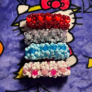 May include: Four colorful beaded bracelets. The top bracelet is red with red heart-shaped beads. The second bracelet is silver with clear beads. The third bracelet is light blue with blue beads. The bottom bracelet is white with pink star-shaped beads.