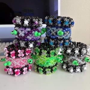 May include: A collection of colorful beaded bracelets. The bracelets are stacked and feature various colors, including pink, purple, blue, green, and black. Some bracelets have star and flower accents, and all have green alien head embellishments.