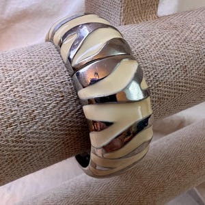 Könnte beinhalten: Ein weißes und silbernes Armreif-Armband mit geometrischem Muster. Das Armband ist aus Metall und hat eine glatte Oberfläche.