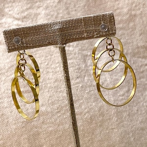 May include: Gold hoop earrings with three interlocking circles. The earrings are on a display stand.