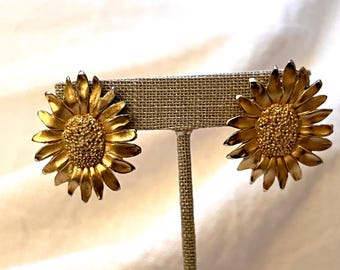 May include: A pair of gold-tone sunflower earrings. The earrings are clip-on style and feature a detailed sunflower design.