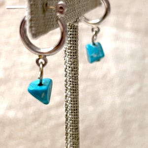 Peut inclure: Une paire de boucles d'oreilles créoles en argent avec des breloques en pierre turquoise. Les boucles d'oreilles sont suspendues à un présentoir.