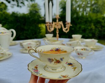 May include: A hand holds a teacup and saucer set. The ivory-colored teacup and saucer are adorned with floral patterns in shades of blue, pink, and yellow, with gold trim. Other teacups, saucers, and a teapot are on a white tablecloth, with a gold candelabra in the background.
