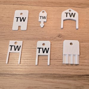 Peut inclure: Divers porte-clés et étiquettes en plastique blanc, chacun avec les lettres "TW" en noir. Les formes varient, incluant des clés, des rectangles et des formes en U. Ils sont disposés sur une surface en bois brun clair.