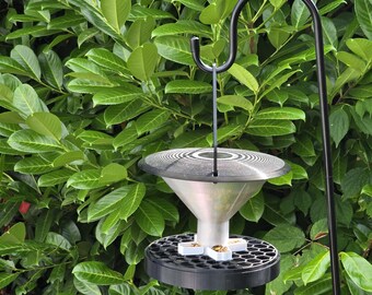 May include: A bird feeder with a black metal hanging hook. The feeder has a silver cone-shaped body, a black base with a honeycomb pattern, and a circular top. The background is a dense green hedge.