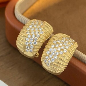 May include: A pair of gold-toned hoop earrings. Each earring features a textured, ridged design and is encrusted with small, sparkling gemstones. The earrings are displayed on a brown surface.