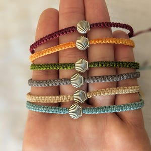 Può includere: Una collezione di braccialetti in macramè colorati, ognuno con un ciondolo a forma di conchiglia argentata. I braccialetti sono in vari colori, tra cui bordeaux, arancione, verde, grigio, beige e turchese. I braccialetti sono esposti su una mano.