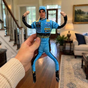May include: A doll of a race car driver in a light blue uniform with black accents. The doll has a blue cap and the word "Telefonica" is printed on the front. The doll is being held up by a person.
