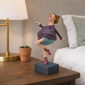 May include: A quirky figurine depicting a person in a dancing pose. The figure is dressed in a purple top, blue shorts, and red socks with white polka dots. It stands on a dark blue rectangular base, placed on a wooden surface. A lamp and bed are in the background.