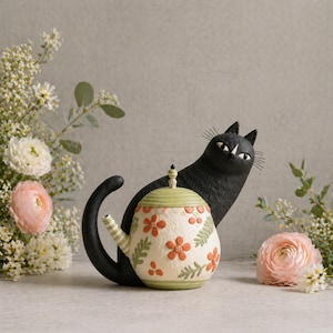 Könnte beinhalten: Eine skurrile Keramik-Teekanne mit einem Blumenmuster in Orange und Grün, begleitet von einer schwarzen Katzenfigur. Die Teekanne hat eine cremefarbene Basis mit orangefarbenen Blumen und grünen Blättern. Die Katze ist schwarz mit weißen Akzenten.