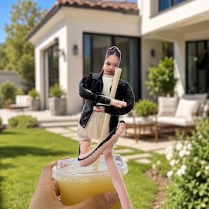 Puede incluir: Una mano sostiene un vaso de plástico transparente lleno de líquido amarillo. Una pajita decorativa con una figura de una persona se inserta en la bebida. La figura lleva una chaqueta negra y un vestido blanco. El fondo muestra una casa y un césped verde.