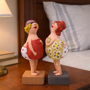 Può includere: Due fantasiose statuette in ceramica di donne, ciascuna in piedi su una base quadrata. Una indossa un asciugamano rosso e un copricapo fantasia, l'altra un indumento giallo a fantasia floreale. Entrambe hanno tratti esagerati e guardano in alto.