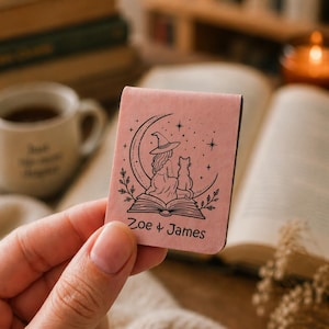 May include: A rectangular pink magnetic bookmark featuring a black line art illustration of a person in a witch hat sitting with a cat on an open book under a crescent moon and stars, with the text Zoe & James printed at the bottom.