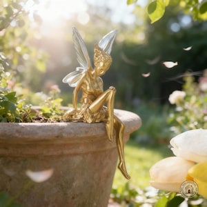 May include: A gold-coloured fairy ornament with clear wings sits on a terracotta plant pot. The fairy is in a seated position, with one leg extended. The background is a blurred garden scene with sunlight.