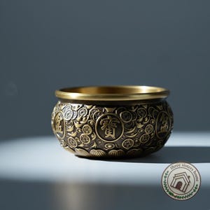 Puede incluir: Un cuenco decorativo redondo de color bronce con intrincados patrones en relieve y caracteres chinos. El cuenco tiene un borde ancho y un diseño detallado de flores y monedas. El cuenco probablemente se utiliza con fines decorativos o como un pequeño recipiente.