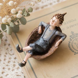 May include: A small figurine of a person with a pointed acorn cap hat, wearing a grey long-sleeved shirt and dark grey overalls, resting inside a brown textured walnut shell. The figure has closed eyes and a relaxed expression with arms outstretched.