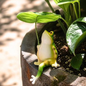 Puede incluir: Una caprichosa figura de rana, pintada en tonos verdes y amarillos, está posada en el borde de una maceta. La rana sostiene una hoja por encima de su cabeza, creando un encantador acento decorativo para un jardín o interior.