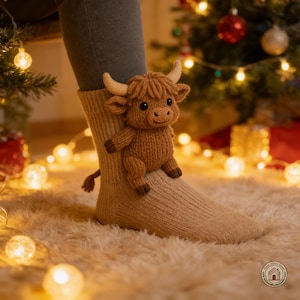 May include: A knitted Highland cow toy in shades of brown perches on a beige sock. The cow features small horns and a fluffy mane. The scene is set in front of a Christmas tree with warm lights, creating a cosy, festive atmosphere.