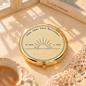 May include: A round, gold-coloured metal compact mirror featuring a black line-art graphic of a rising sun. The text "One sun, two horizons." is printed above the sun, with "M 06:42" and "J 19:18" positioned on the left and right sides.