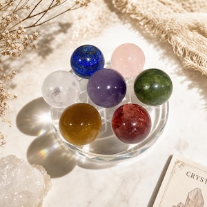 May include: A collection of six polished gemstone spheres arranged in a circular pattern on a clear glass stand. The spheres include a deep blue lapis lazuli, a pale pink rose quartz, a clear quartz, a purple amethyst, a dark green jade, and a golden-brown citrine.