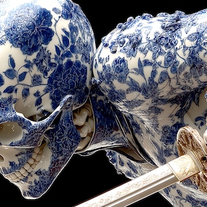 May include: A detailed sculpture of a skull and torso, decorated with a blue and white floral pattern, similar to porcelain. The figure holds a katana with a gold hilt. The artwork is set against a black background.