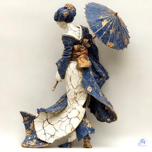 Può includere: Scultura in ceramica di una geisha in kimono tradizionale, con un parasole. Il kimono è blu con dettagli dorati, e la figura ha una base bianca con una texture crepata. Il parasole è anch'esso blu e oro.