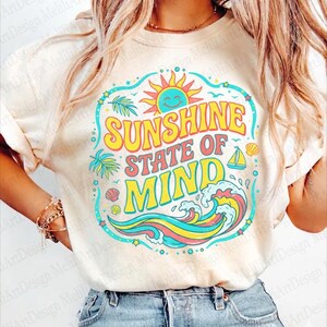 Pode incluir: Camiseta creme com um gráfico colorido que diz "SUNSHINE STATE OF MIND". O design inclui um sol sorridente, ondas, um veleiro e folhas de palmeira. A camiseta é combinada com jeans azuis.