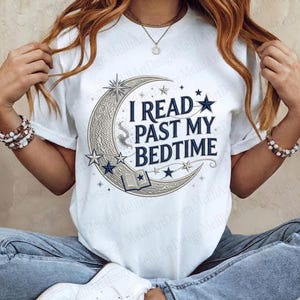 Pode incluir: Camiseta branca com o texto azul marinho "I READ PAST MY BEDTIME", com um design de lua crescente e estrelas. O design inclui um livro aberto e estrelas. Uma peça de roupa casual.