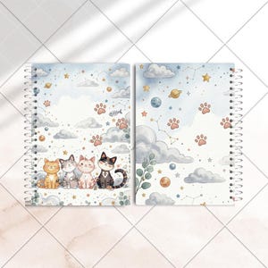 Pode incluir: Dois cadernos com espiral com tema de gato. As capas apresentam ilustrações em aquarela de gatos, pegadas, nuvens, estrelas e planetas. A paleta de cores inclui azuis, laranjas, cinzas e brancos, criando um design celestial e sonhador.
