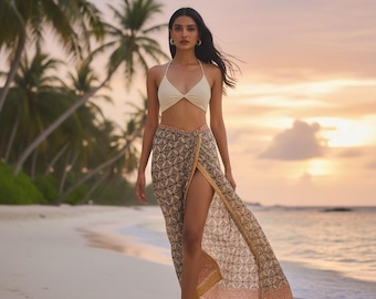 Puede incluir: Una mujer con un top halter color crema y una falda envolvente estampada con una abertura alta, caminando por una playa de arena. La falda presenta un diseño geométrico en marrón, beige y melocotón. Hay palmeras y el océano al fondo.